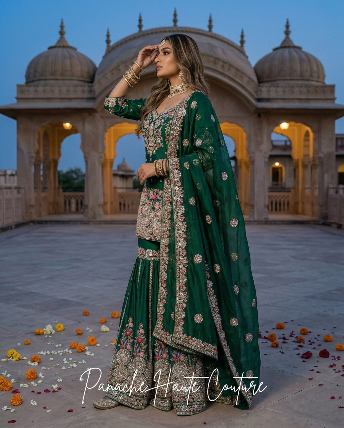 A woman models a traditional Indian green lehenga choli set with intricate gold embroidery, standing in front of a historic domed stone structure at dusk. The brand name 'Panache Haute Couture' is watermarked at the bottom.